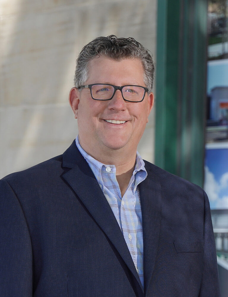 A smiling man in glasses, a button-down shirt, and a navy blazer.