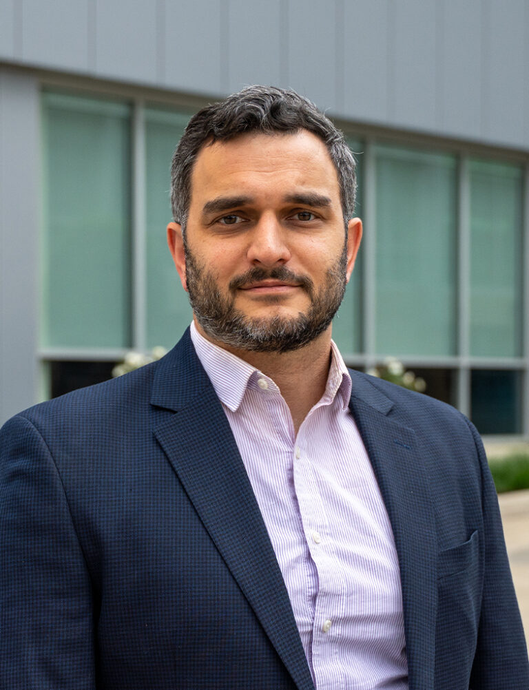 A man with a beard wearing a navy blue blazer and a light-colored button-up shirt.