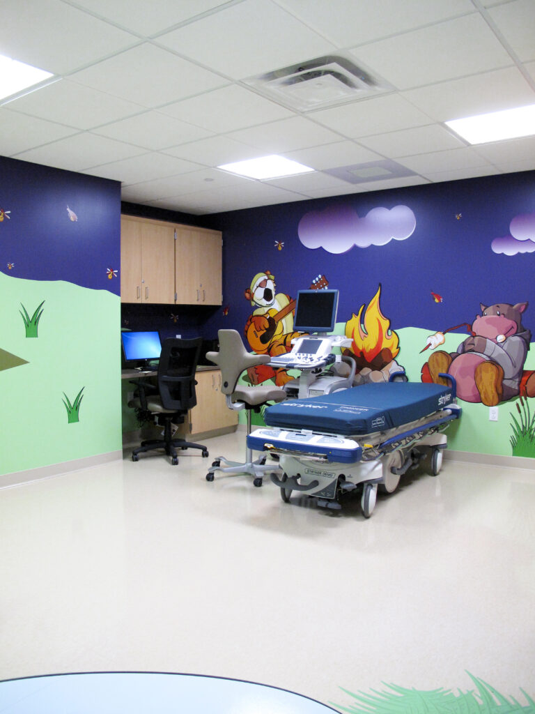 Medical exam room featuring a gurney and ultrasound machine against a wall decorated with a cartoon mural of animals camping.