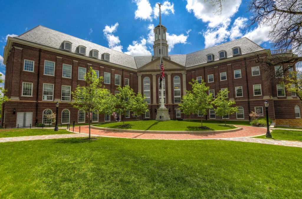 A multi-story brick academic building with a central cupola, flag pole, and a curved brick walkway surrounded by green lawns and trees.
