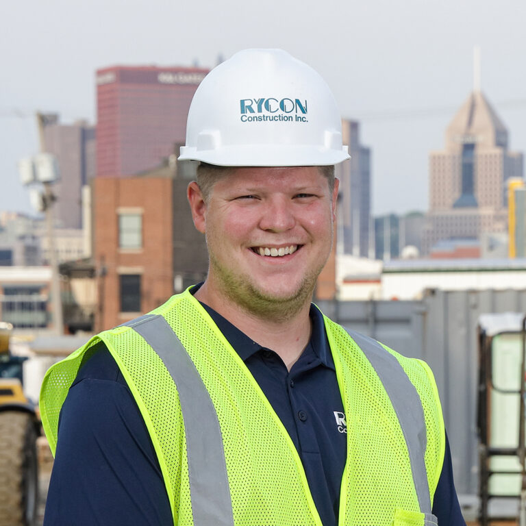 A smiling construction worker wearing a white Rycon Construction hard hat and a high-visibility safety vest, with a city skyline in the background.
