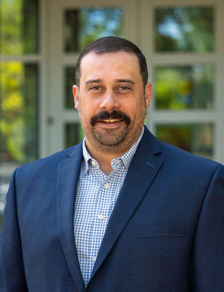 A man with a mustache wearing a blue plaid shirt and a navy blazer.