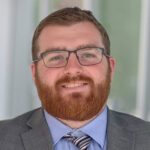 Headshot of a man with a beard and glasses wearing a suit, dress shirt, and tie.