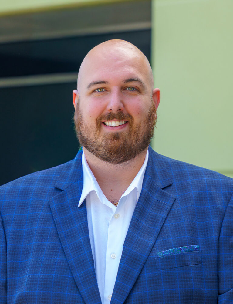 A smiling man with a shaved head and a beard, wearing a blue plaid blazer over a white dress shirt.