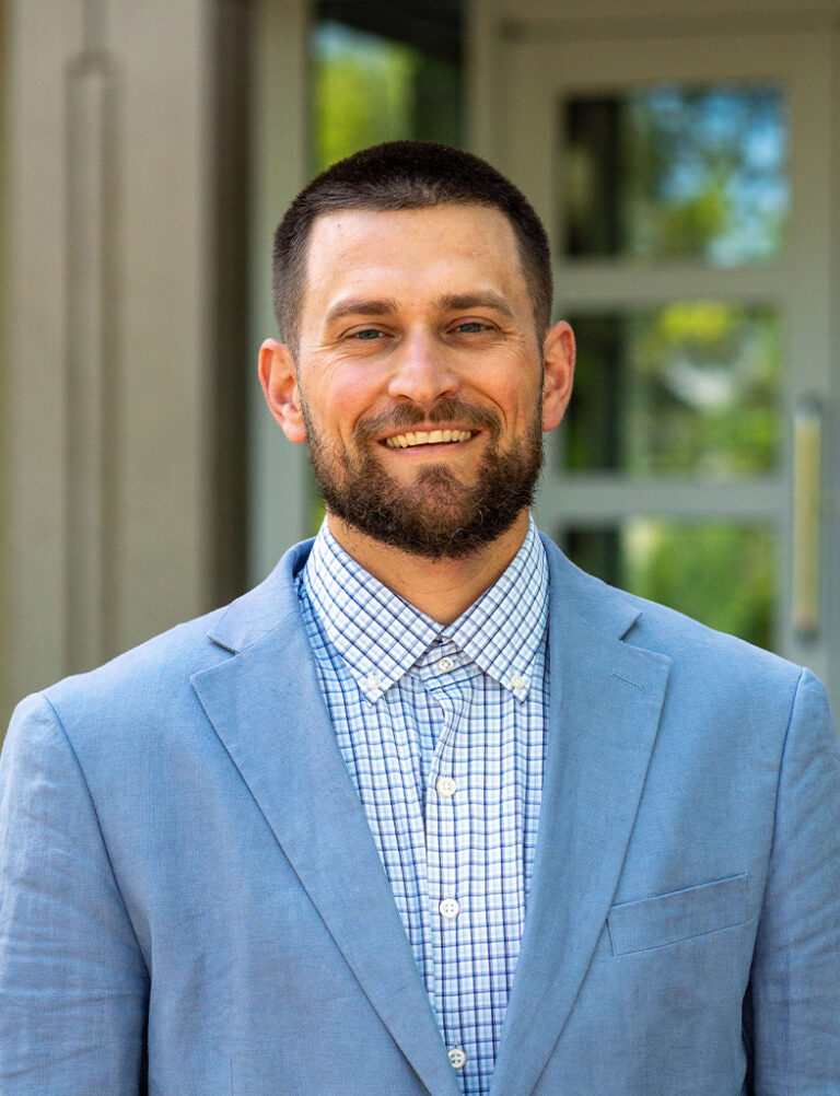 A smiling man with a short beard wearing a light blue blazer over a blue-and-white checkered button-down shirt.