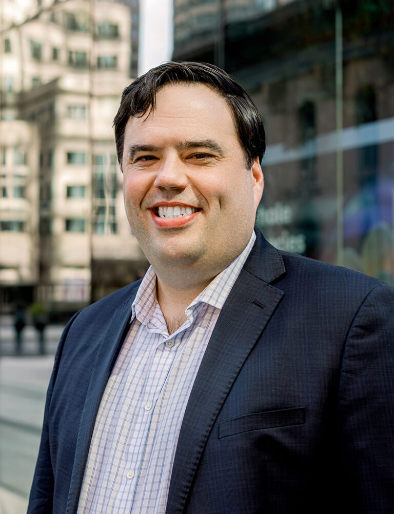 A man with dark hair smiling, wearing a blue plaid shirt under a dark blazer.
