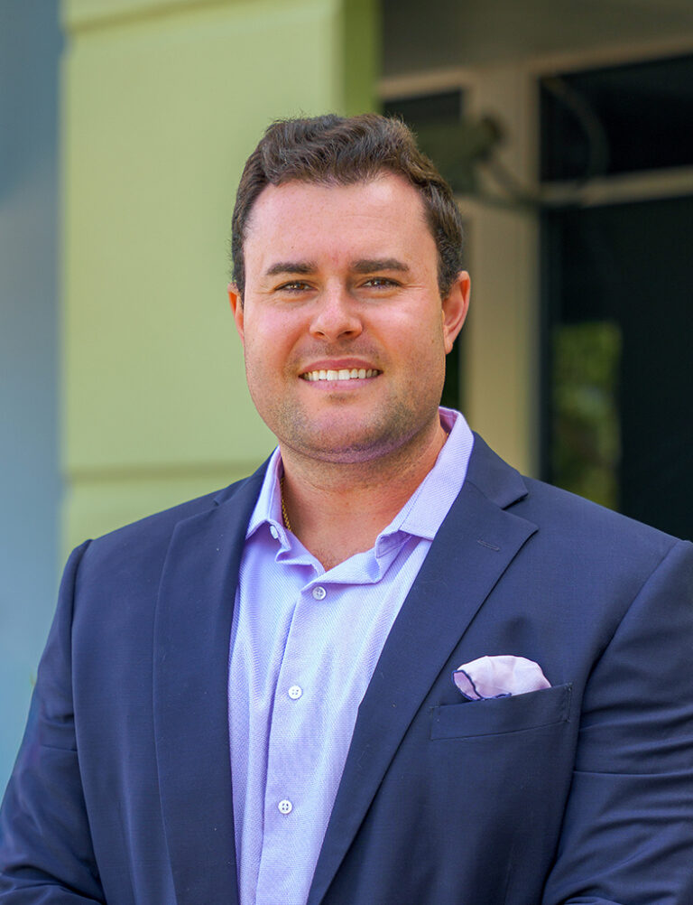 Headshot of a smiling man in a navy blue blazer and light purple button-down shirt with a pocket square.