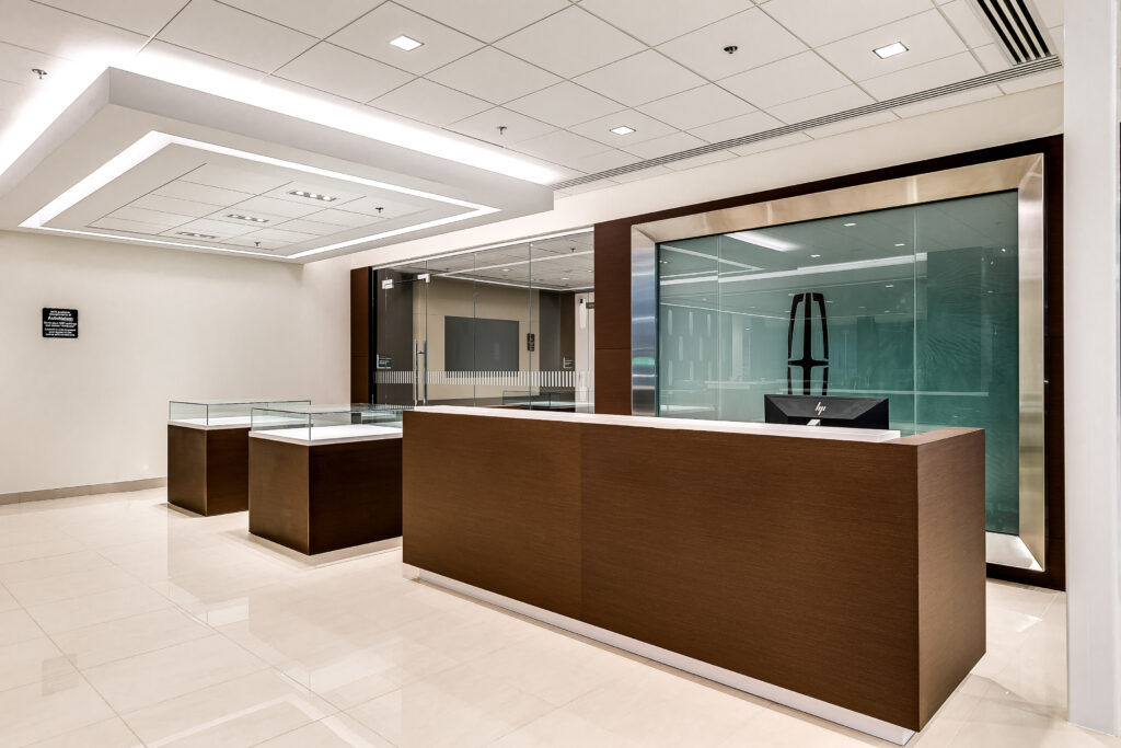 Reception area with a long, dark wood desk and two glass display cases on a light-colored tile floor, featuring a Lincoln logo on a glass partition in the background.