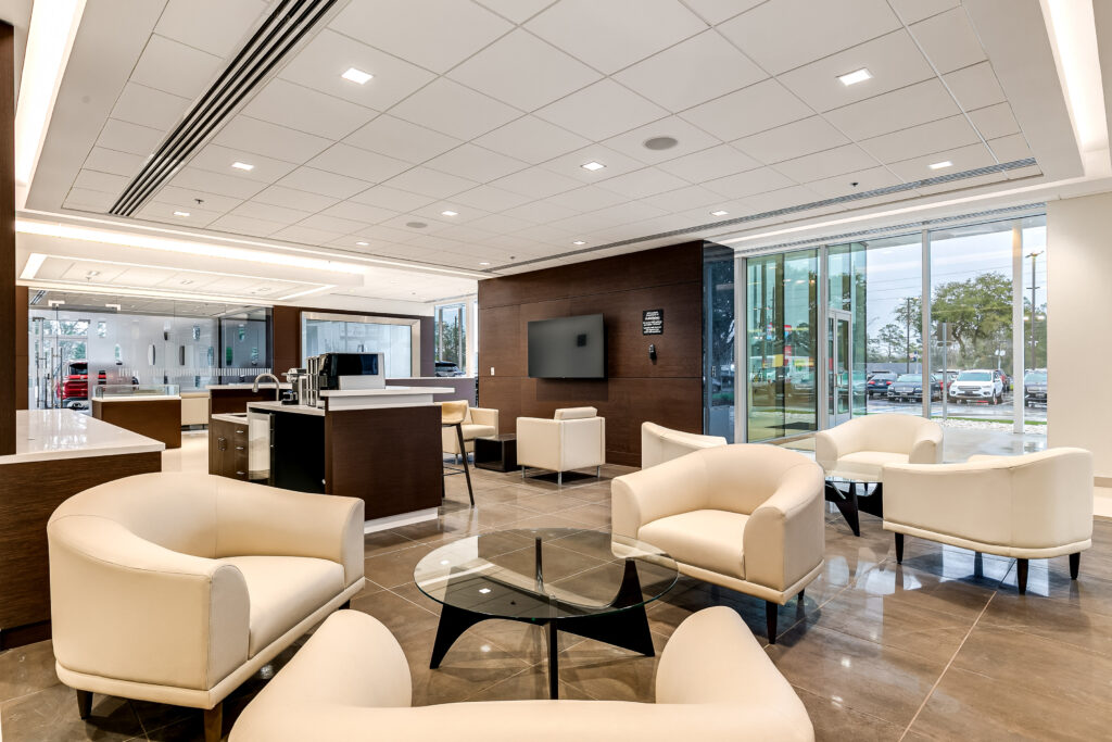 A modern dealership lounge with cream-colored armchairs, a glass coffee table, and a service counter in front of floor-to-ceiling windows.