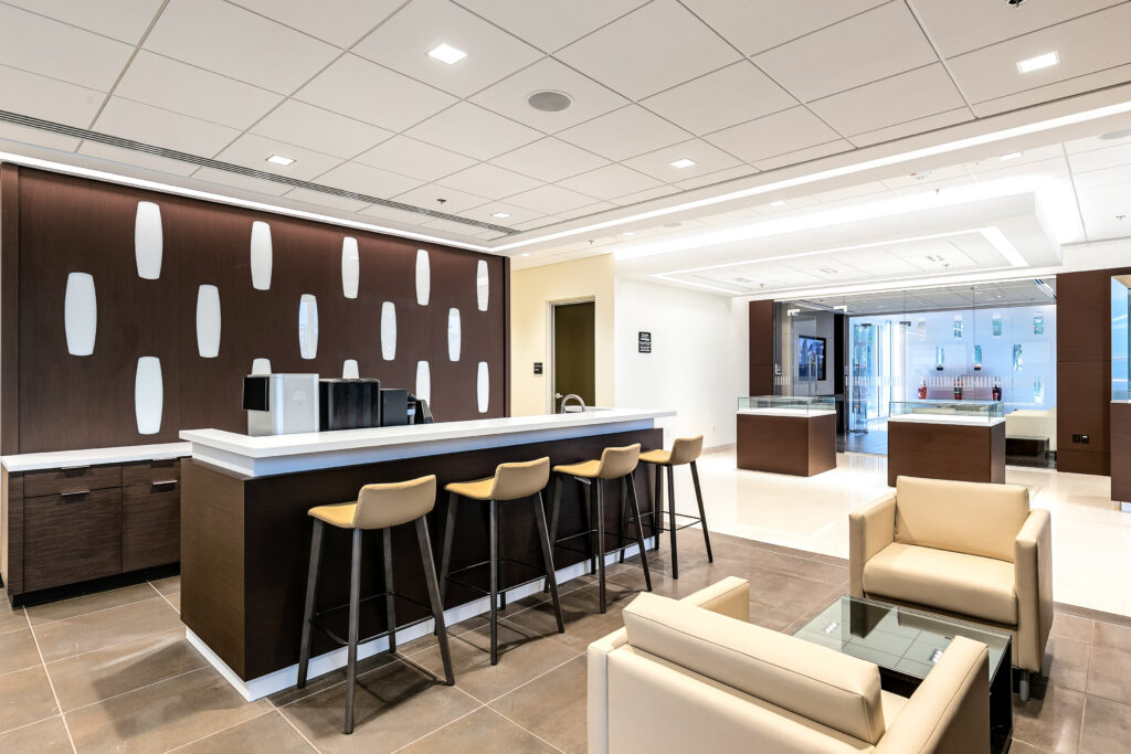 Modern office breakroom featuring a dark wood counter with four bar stools, facing two beige lounge chairs and a glass coffee table.