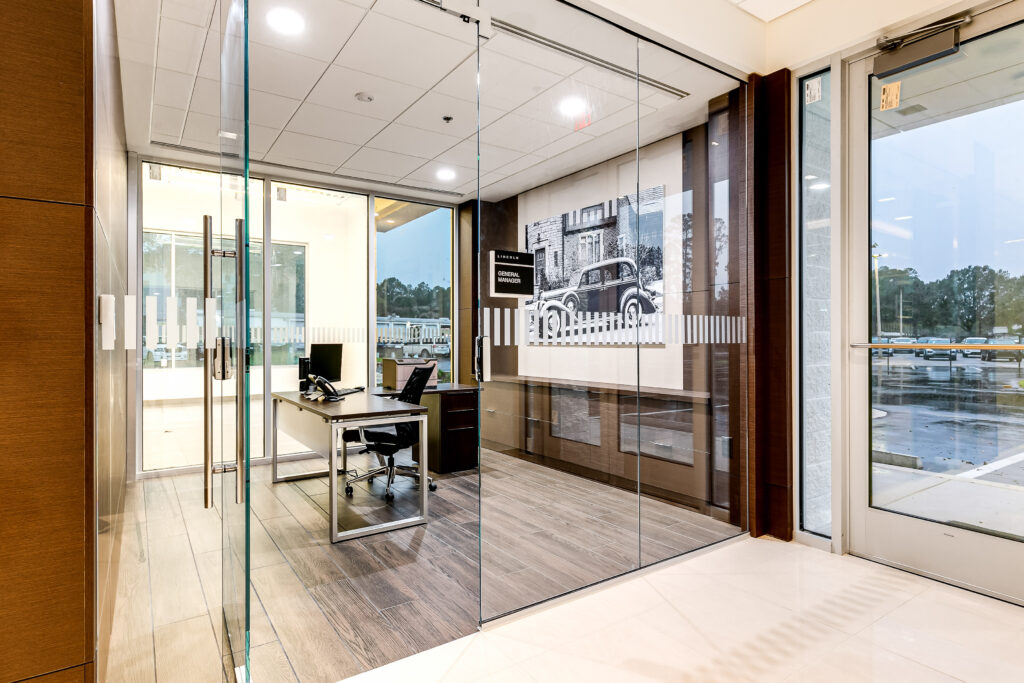 A modern office enclosed by glass walls, featuring a desk, rolling chair, and a framed black-and-white photograph of a vintage car on the wall.