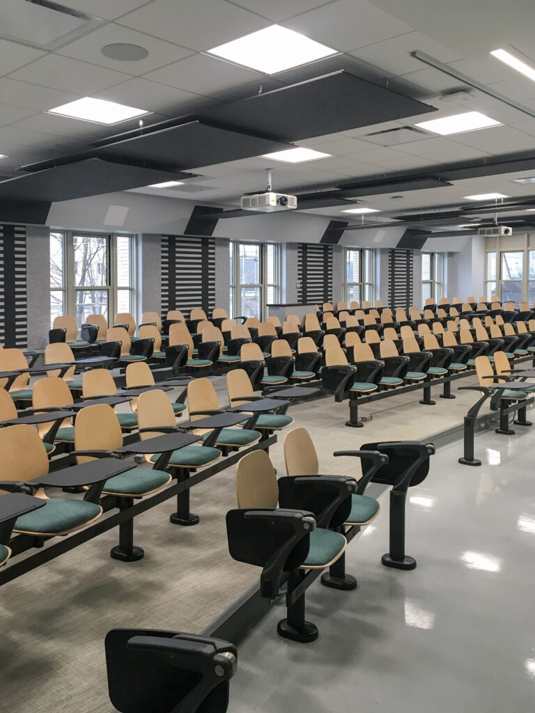 Rows of tiered lecture hall seating with light wood backs and teal cushioned seats, each featuring an attached writing desk.