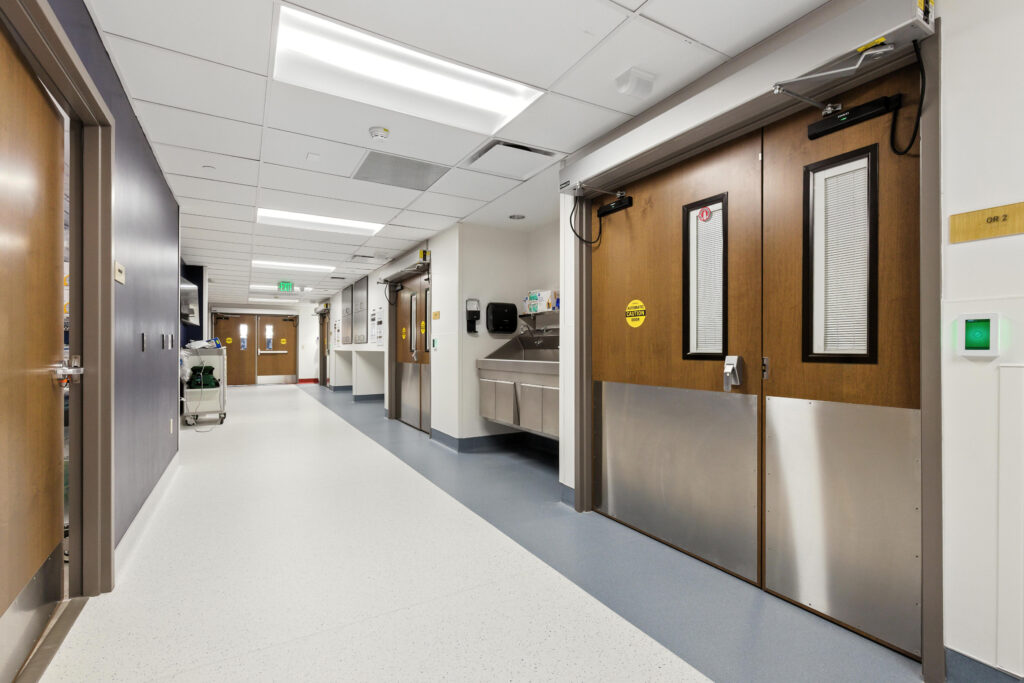 A bright, long hospital corridor with double wooden doors, a metal sink station, and linoleum floors.