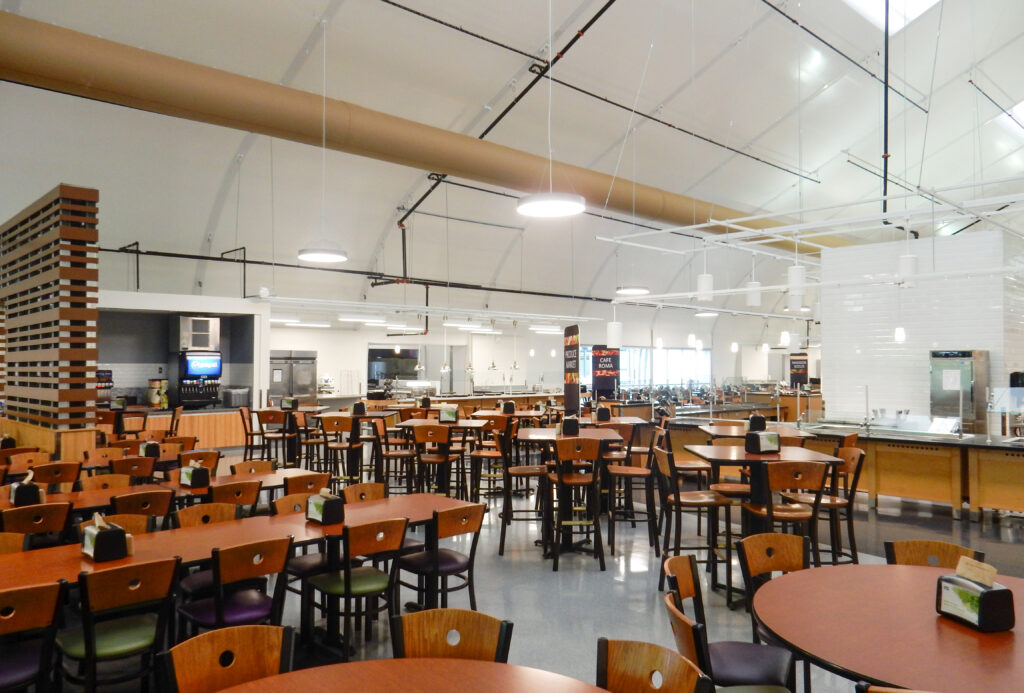 Large, empty cafeteria with rows of tables and chairs, stainless steel serving counters, and a beverage station.