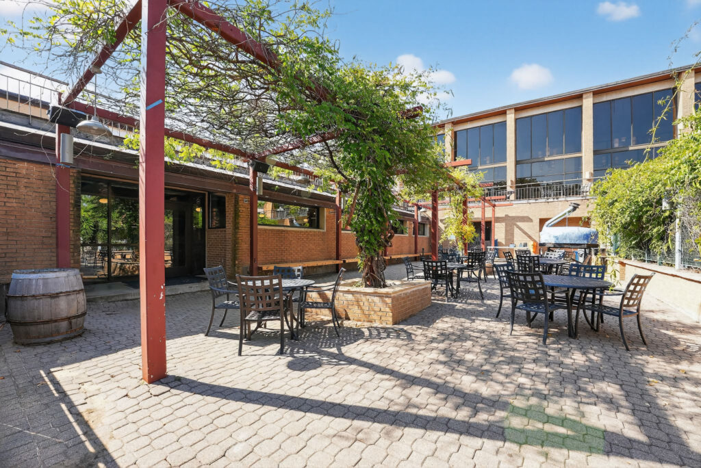 Outdoor patio with metal tables and chairs under a vine-covered pergola in front of a brick building.