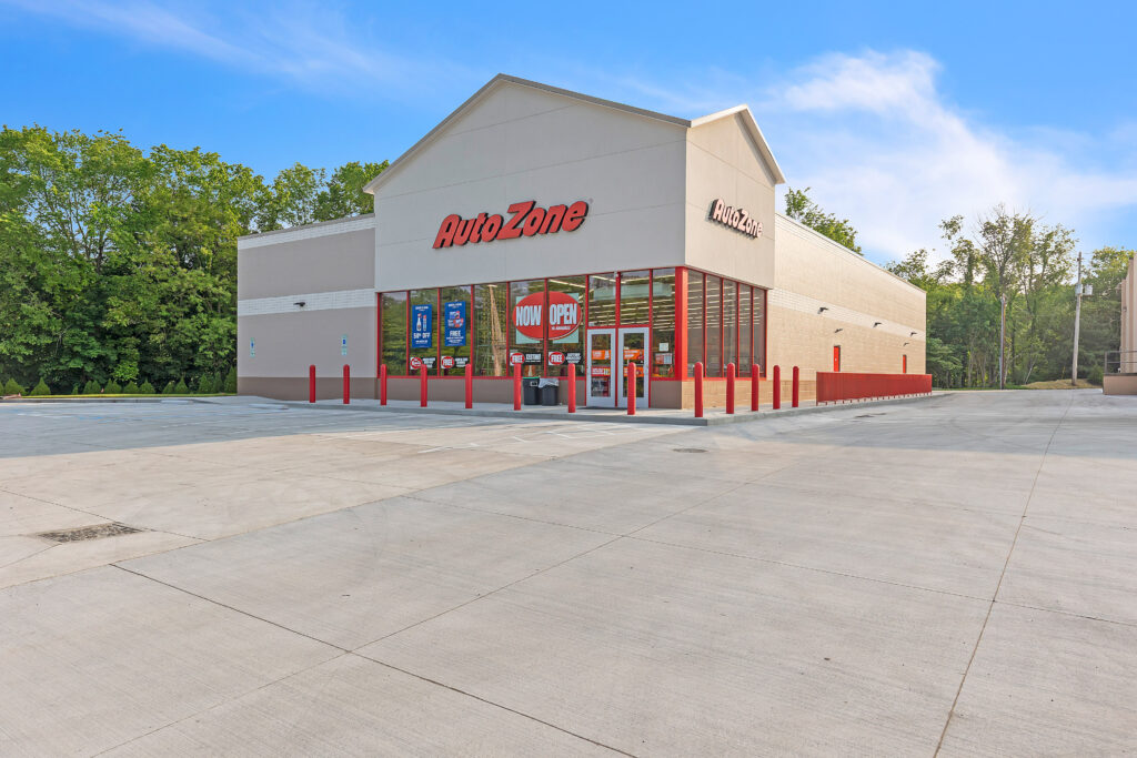 An AutoZone store exterior with a “Now Open” sign in the front window and an empty concrete parking lot.