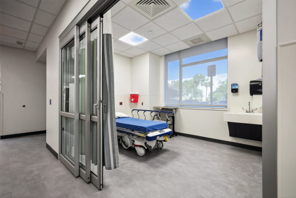 A hospital room with a patient bed, a sink, and a large window looking out to palm trees.