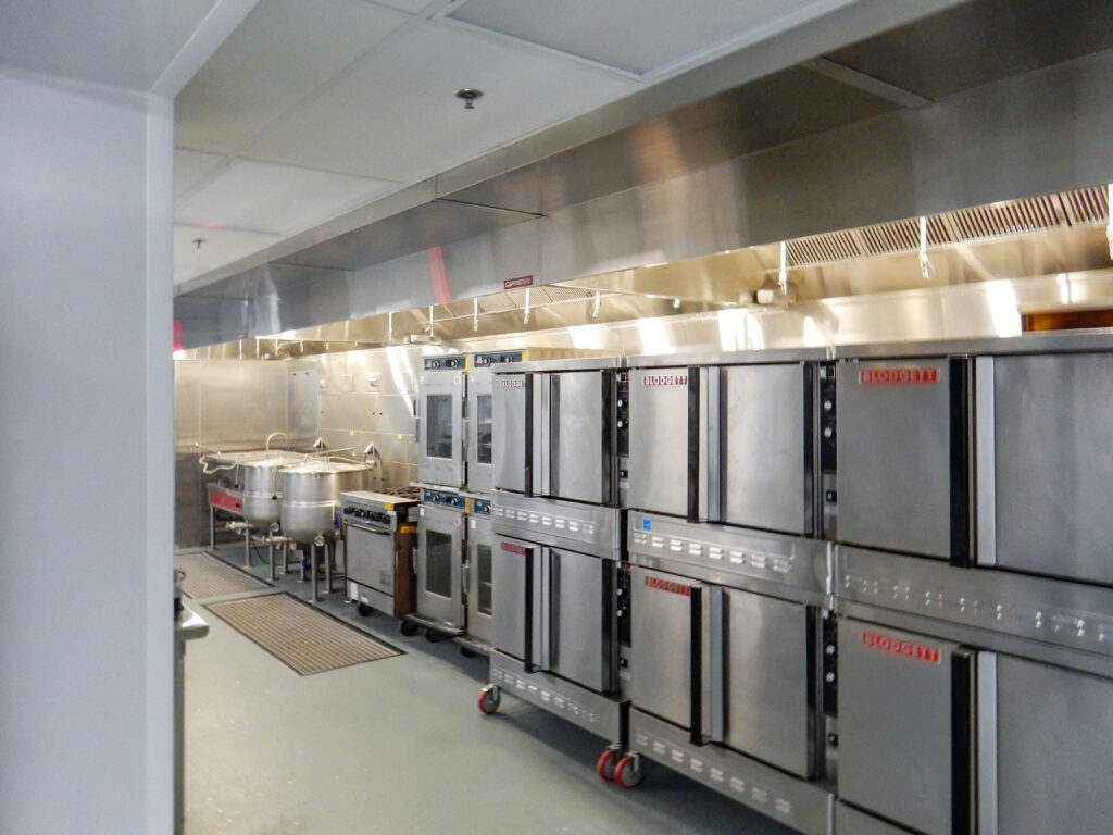 A commercial kitchen featuring rows of stainless steel convection ovens and large stockpots under a metal vent hood.