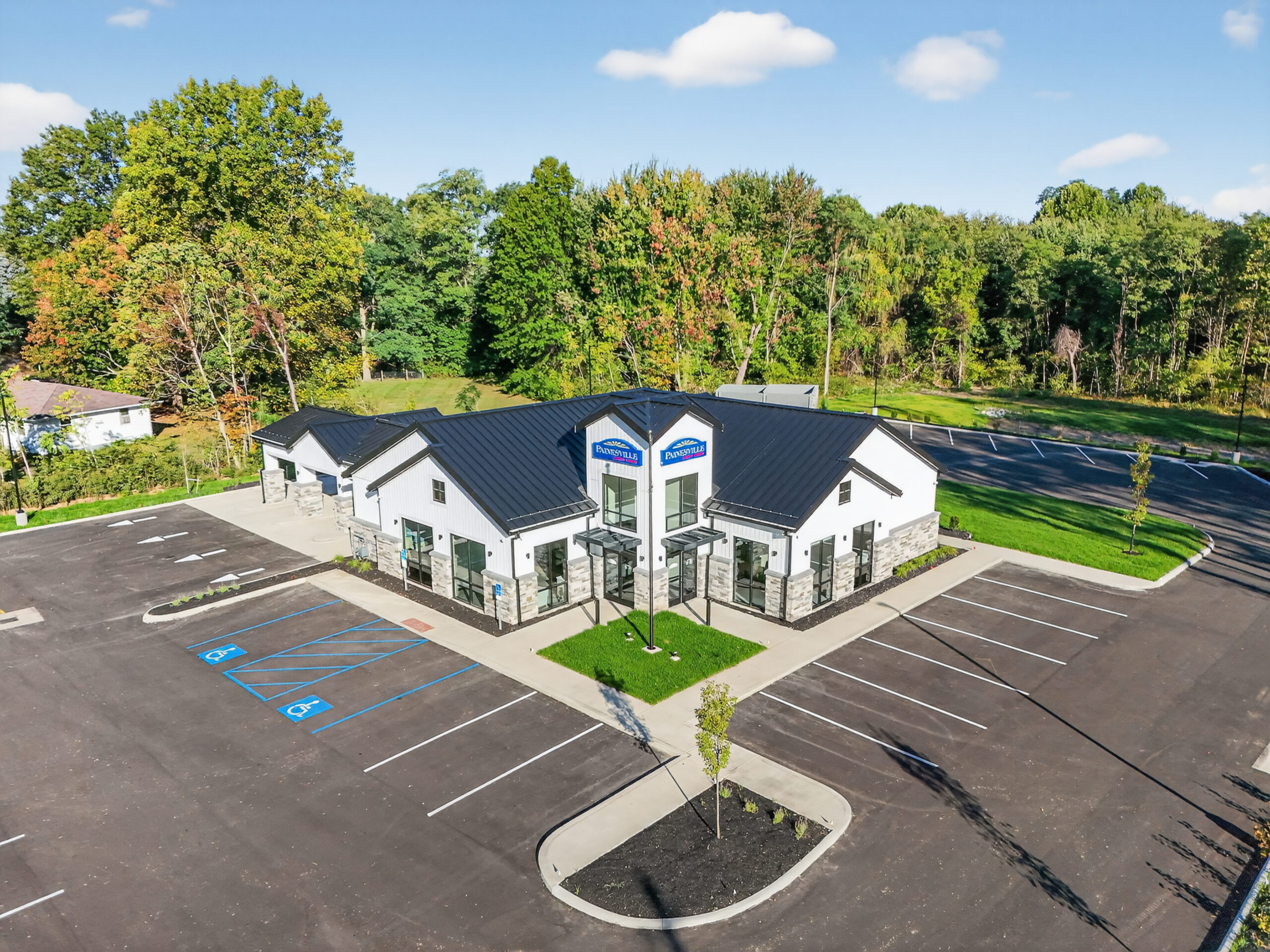 Aerial view of a white commercial building with a black metal roof and “Painesville” signage, surrounded by a paved parking lot and trees.