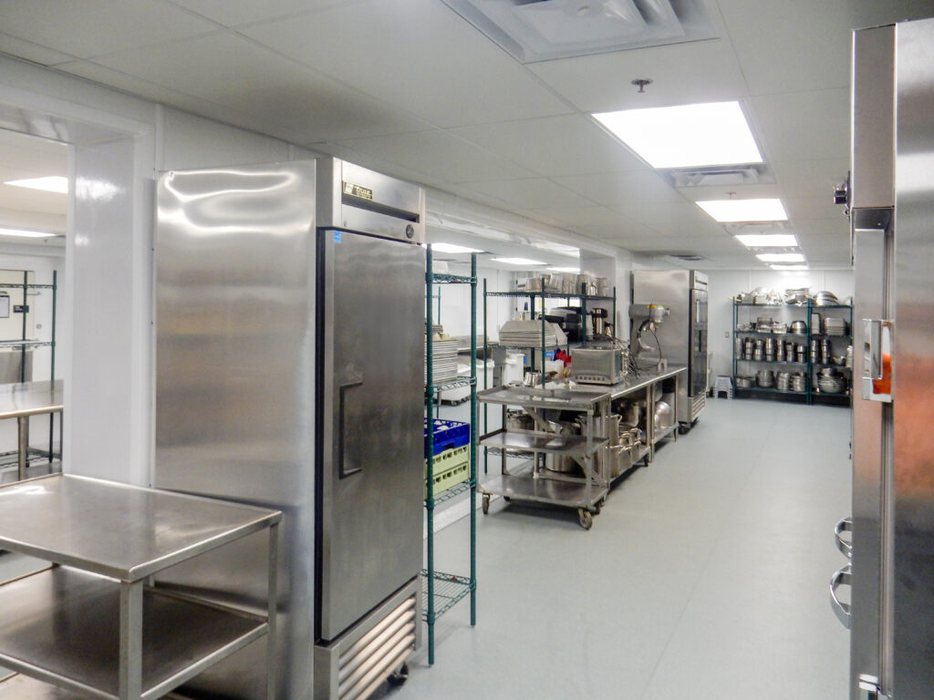 Commercial kitchen featuring stainless steel refrigerators, prep tables, wire shelving units, and various cookware.