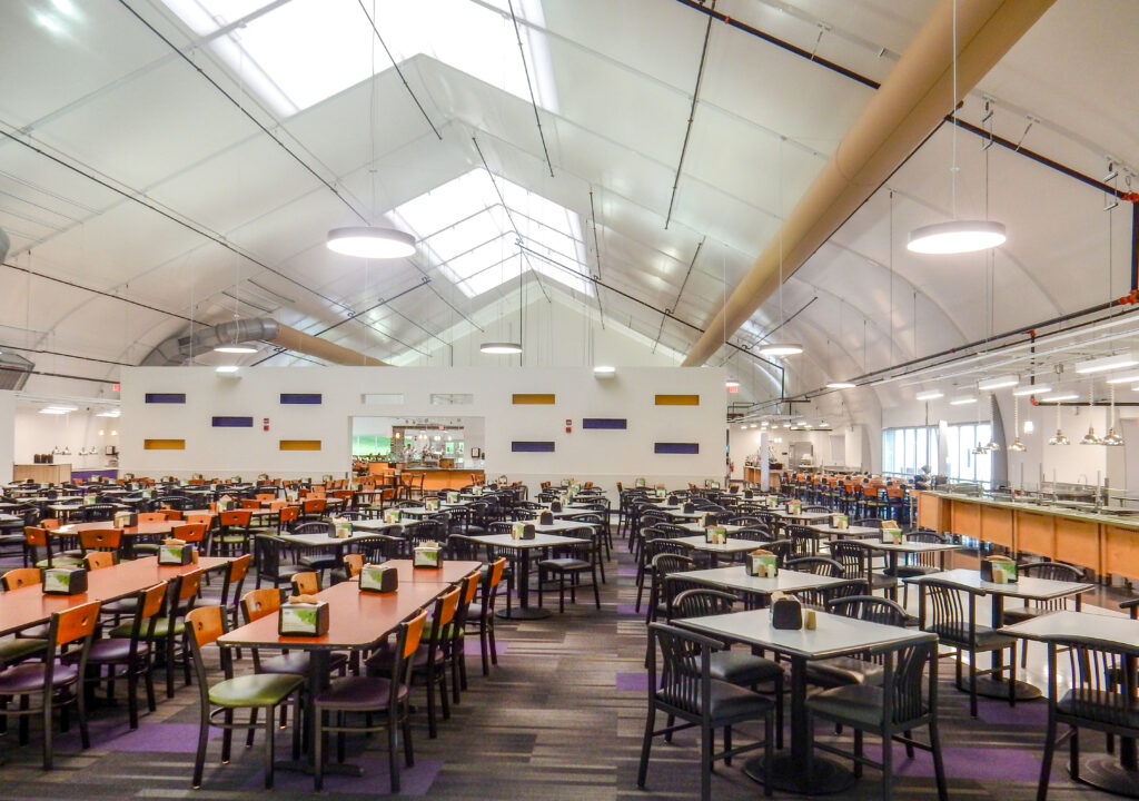 A large, open-plan dining hall featuring numerous tables with chairs and a serving counter in the background, set under a high-arched ceiling with skylights.