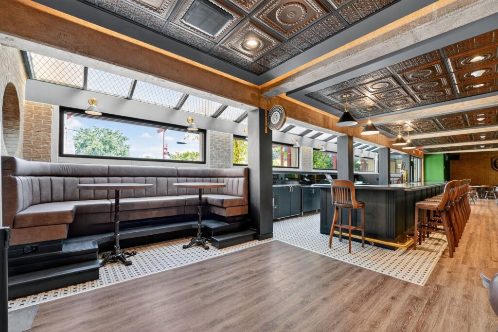 Interior view of a restaurant featuring a long grey booth, small circular tables, and a bar with wooden stools beneath a decorative tin ceiling.