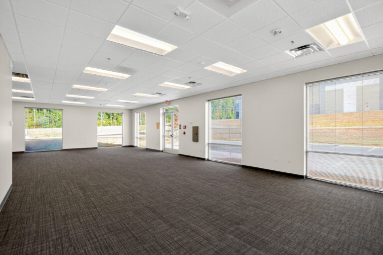 An empty commercial office space with gray carpet, white walls, drop ceiling, and multiple large windows.