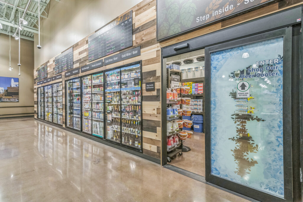 A refrigerated beer section in a store with glass-door coolers and a walk-in cooler entrance featuring a sign that reads, “Enter Shop Our Beer Cooler.”
