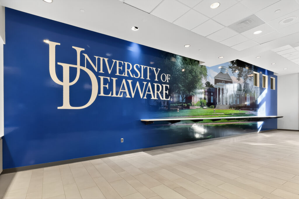 A blue wall featuring the University of Delaware logo and a photographic mural of campus, with a shelf mounted along the wall.