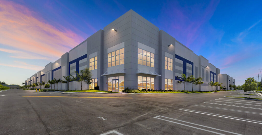 A modern commercial warehouse building at dusk, featuring a large parking lot and glass-paneled corner entrance.