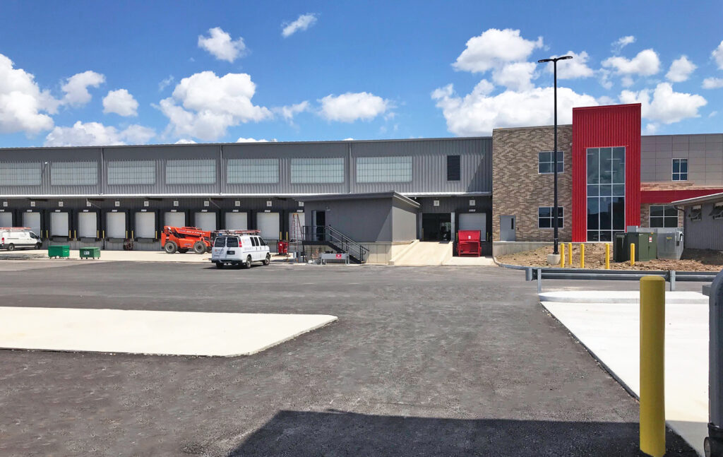 Large industrial warehouse building featuring multiple loading docks, a parking lot, and a red accent wall at the entrance.