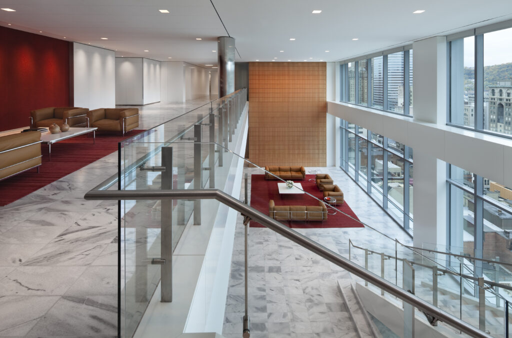 Two levels of a modern office lobby connected by a glass-railed staircase, featuring lounge seating areas on marble floors and floor-to-ceiling windows overlooking a city.