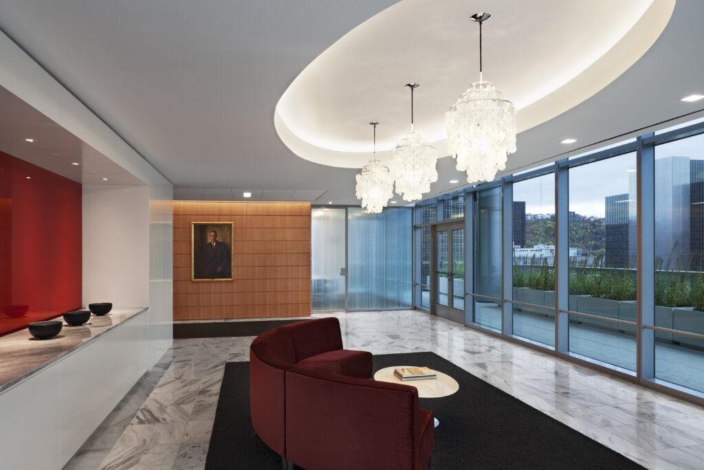 Lobby featuring a curved red sofa, a small circular table, three tiered crystal chandeliers, and a portrait hanging on a wood-paneled wall.
