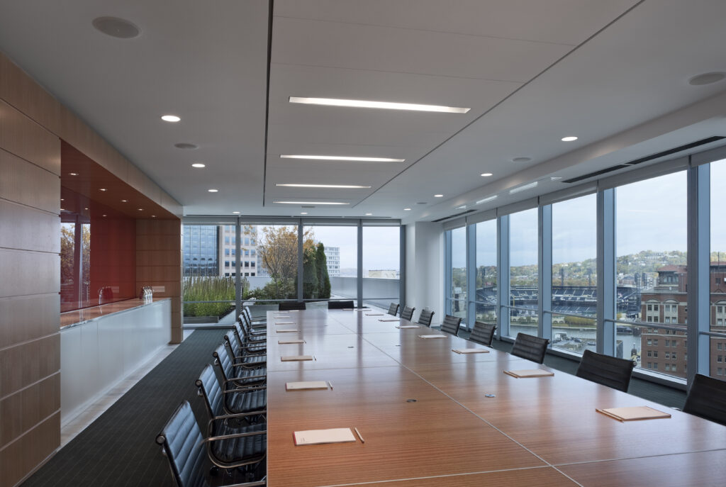 A long wooden conference table surrounded by black chairs in a modern office with floor-to-ceiling windows overlooking a city and stadium.