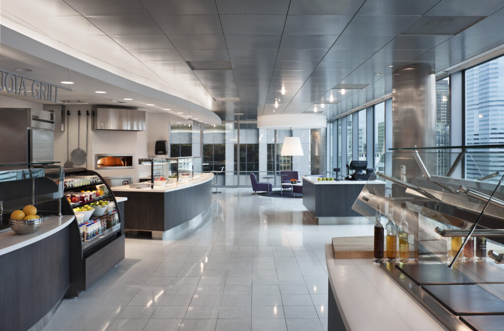 A modern corporate cafeteria with a glass-fronted refrigerated food display, serving counters, and seating areas by large windows.