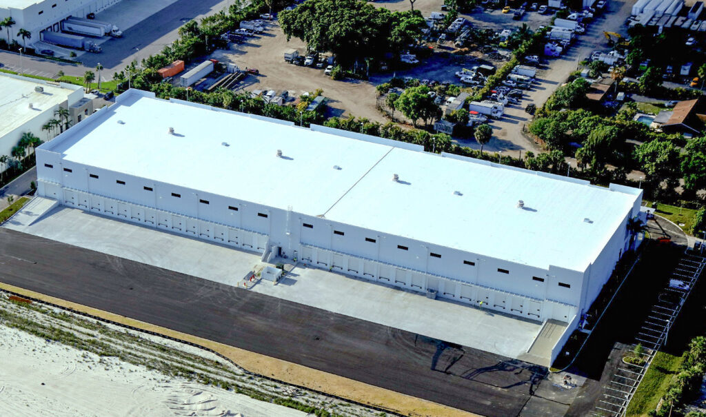 Aerial view of a large white industrial warehouse with multiple loading docks, adjacent to a paved lot and wooded area.