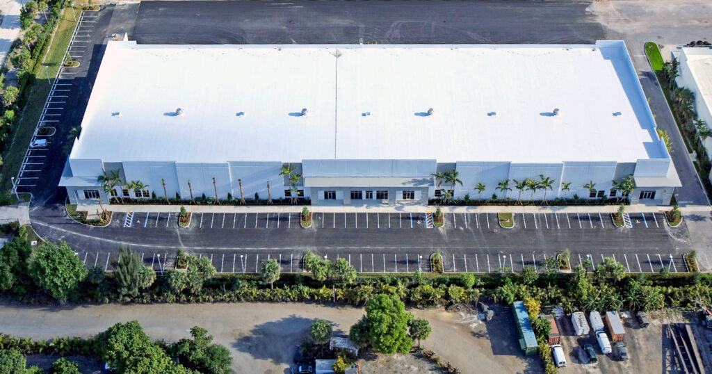 Aerial view of a large rectangular warehouse with a white roof and a long parking lot in front.