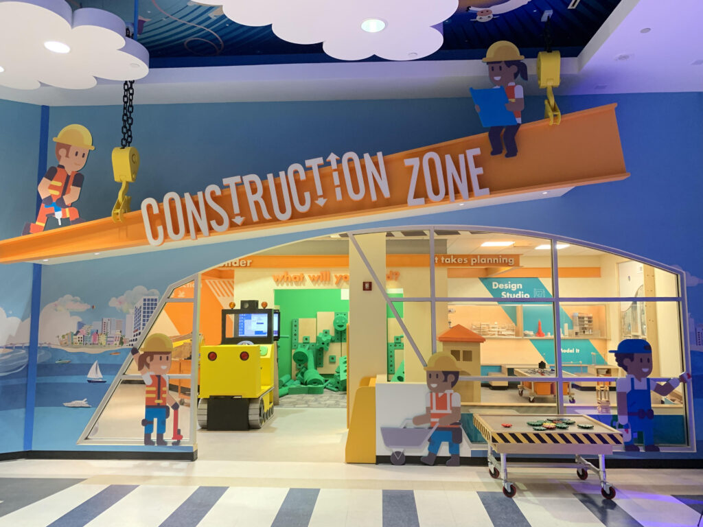 The entrance to a children’s play area themed as a construction zone, featuring colorful cartoon character cutouts, a toy tractor, and building materials.