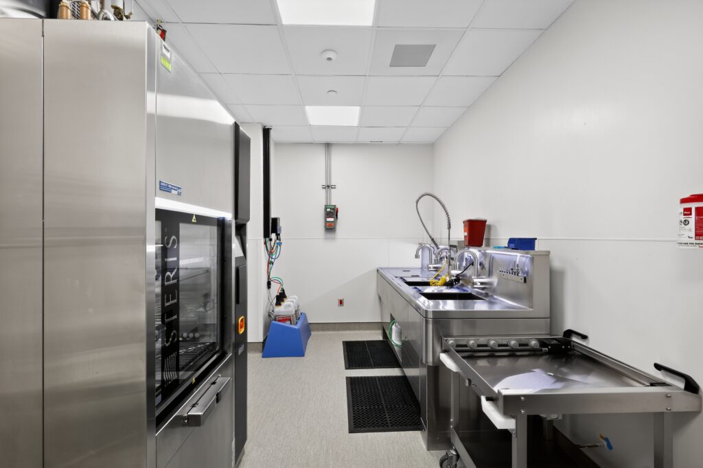 A sterile processing room with a large metal autoclave, a stainless steel sink station with faucets and hoses, and a mobile equipment cart.