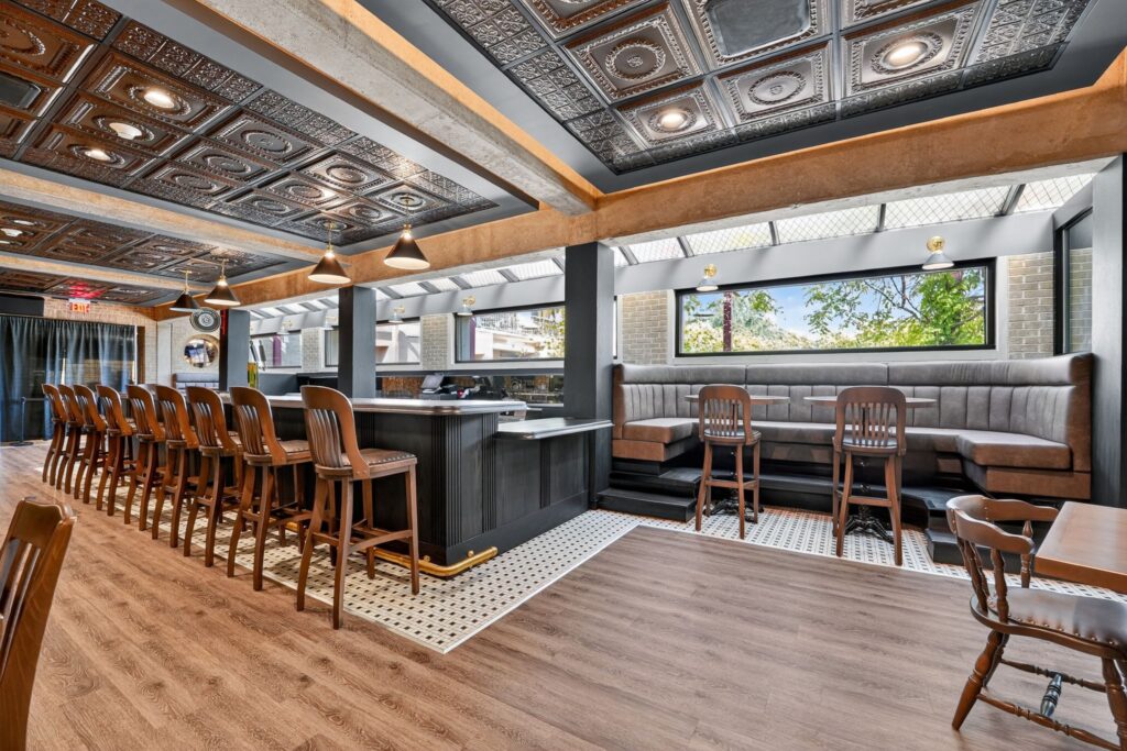 A bar interior featuring a long wooden counter with stools, a corner booth with upholstered seating, and a patterned tin ceiling.