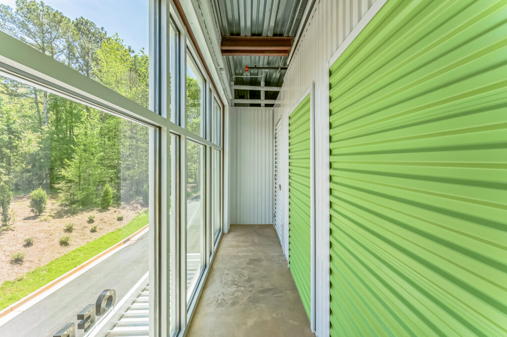 A narrow hallway lined with lime green corrugated metal storage unit doors, beside a wall of floor-to-ceiling windows overlooking trees.