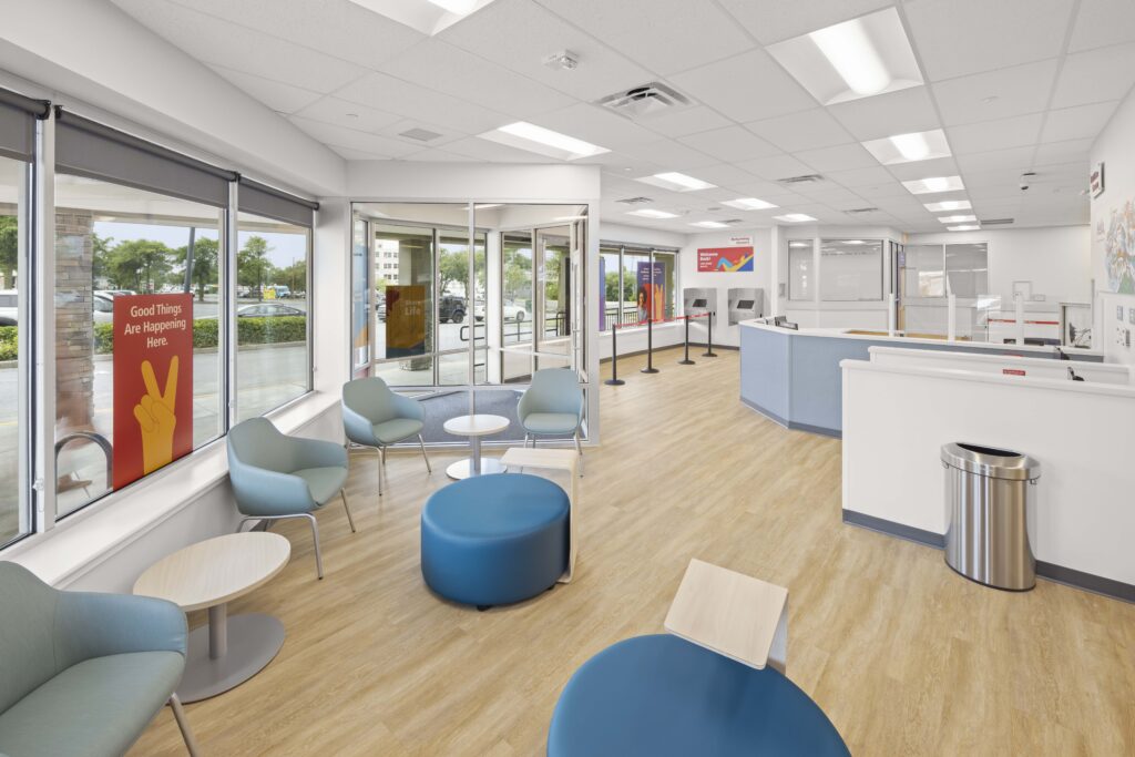 Waiting area of a modern bank office with chairs, blue ottomans, small tables, and a service counter.