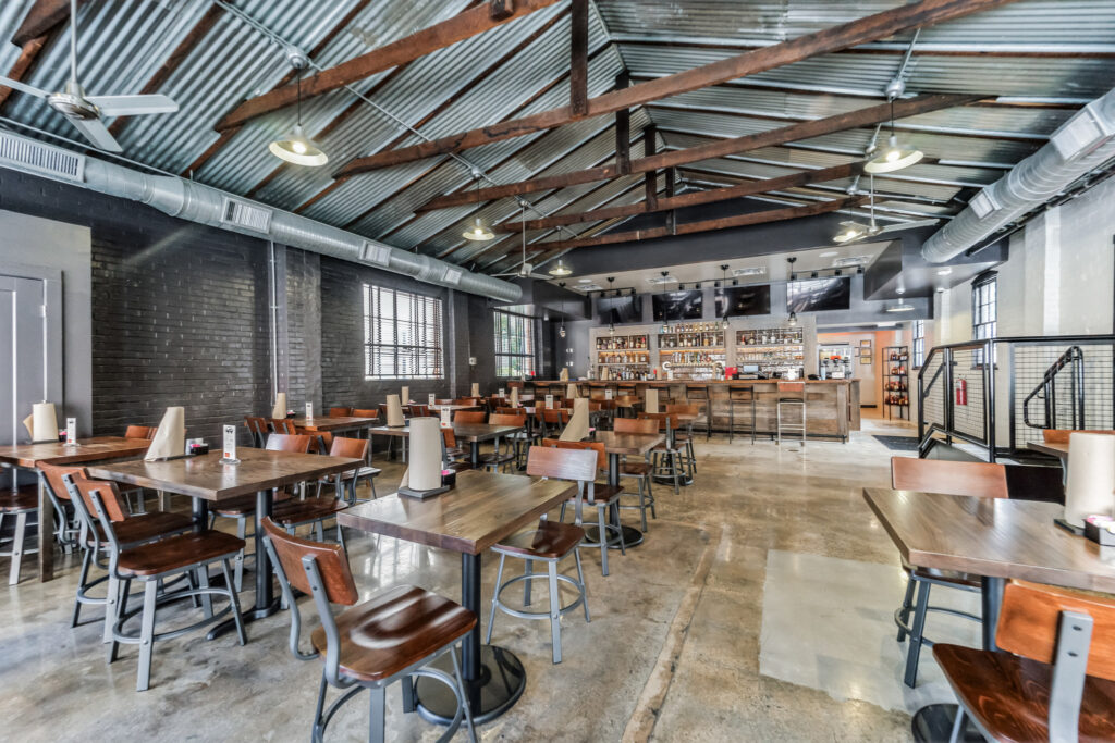 Interior of a restaurant with a high vaulted metal roof, dark brick walls, multiple wooden tables with chairs, and a bar area with stools in the background.