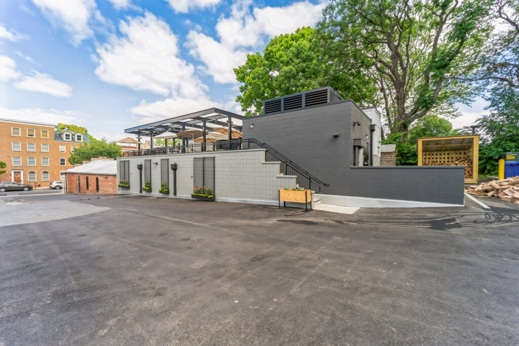A two-story grey building with a rooftop patio, accessed by an exterior staircase, situated next to a parking lot.