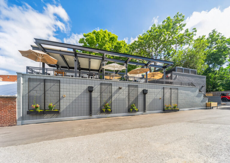 Outdoor dining terrace on a raised grey tiled patio with umbrellas, planters, and a black metal pergola.
