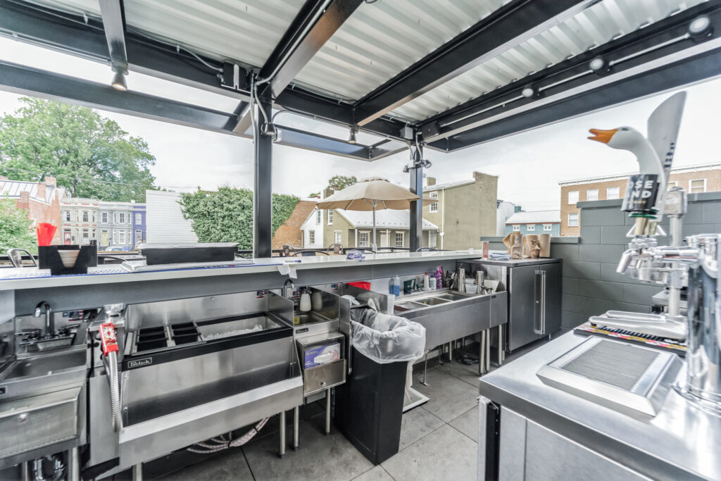 An outdoor bar area featuring stainless steel sinks, a trash bin, and a beer tap with a duck-shaped handle.