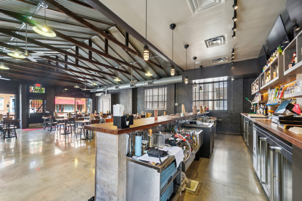 Interior of a restaurant showing a bar counter in the foreground, dining tables in the background, and industrial-style exposed metal ceiling trusses.