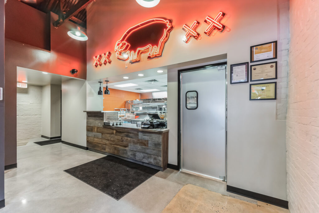 Service counter and kitchen pass-through beneath a neon pig sign, next to a stainless steel door and framed certificates on the wall.