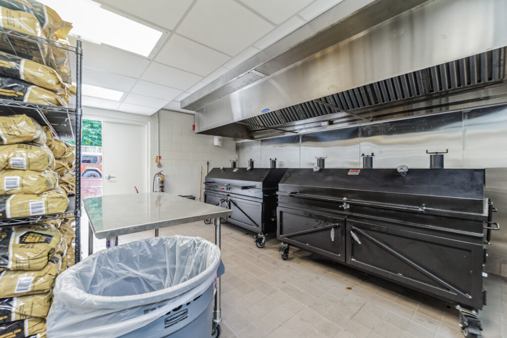 Commercial kitchen featuring two large black smokers under a stainless steel ventilation hood, a metal prep table, and a rack of bagged supplies.