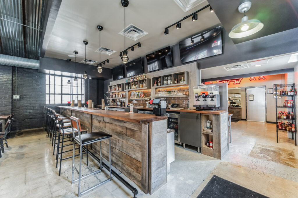 A bar interior featuring a wooden counter with stools, a back bar with shelves of liquor bottles, draft beer taps, and a neon sign in the background.